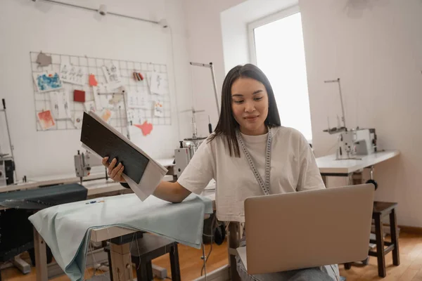 Asiatischer Designer mit Klemmbrett und Laptop im Studio — Stockfoto