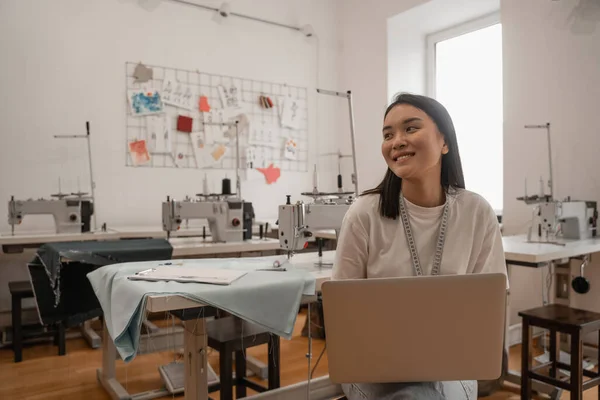 Sorridente designer asiatico con metro a nastro e laptop in atelier — Foto stock