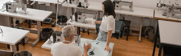 Vista de ángulo alto de diseñador asiático sosteniendo taza de papel cerca de colega y máquinas de coser, pancarta - foto de stock