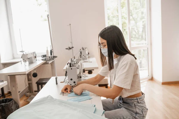 Asian designer in medical mask sewing in atelier — Stock Photo