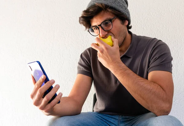 young man student sitting eating an apple and using a phone over white background