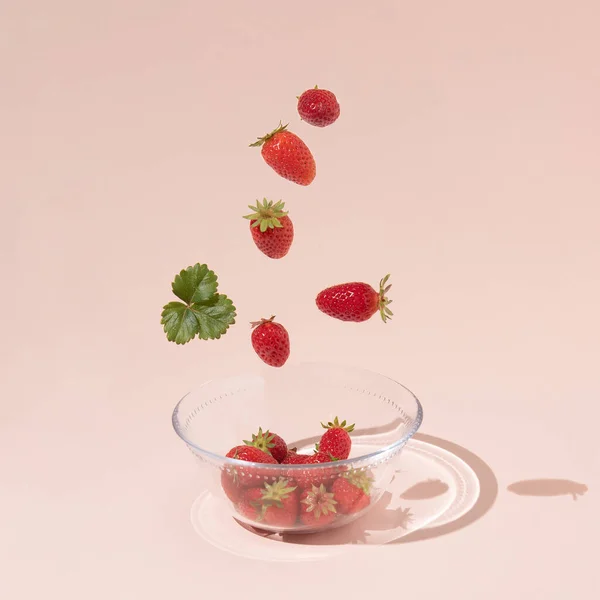 Frutas frescas de verano, fresas y hojas verdes caen en un tazón de vidrio, aisladas sobre un fondo rosa. — Foto de Stock