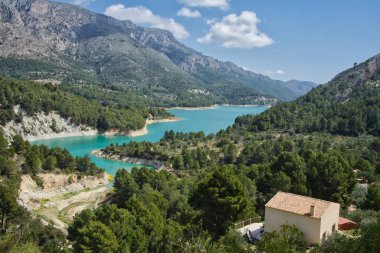Dağlar arasında ağaçlarla çevrili turkuaz bir gölün muhteşem ve güneşli manzarası. İspanya, Alicante 'deki Guadalest rezervuarı.