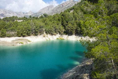 Dağlar arasında ağaçlarla çevrili turkuaz bir gölün muhteşem ve güneşli manzarası. İspanya, Alicante 'deki Guadalest rezervuarı.