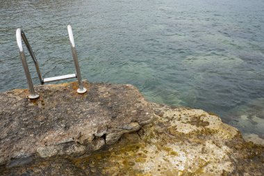 Doğal havuzlar. Metal merdiven sahildeki kayalıklardan denize erişim sağlıyor. Arka plandaki dağlarda üslerinde binalar var. Andrag, Benisa, Alicante, İspanya