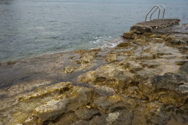 Doğal havuzlar. Metal merdiven sahildeki kayalıklardan denize erişim sağlıyor. Arka plandaki dağlarda üslerinde binalar var. Andrag, Benisa, Alicante, İspanya