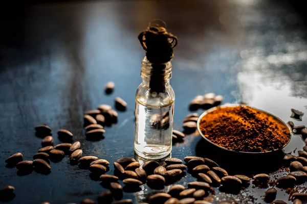 Coffee face mask for anti-aging on a black colored wooden surface consisting of some coffee beans and raw organic coconut oil in a small tiny glass bottle.