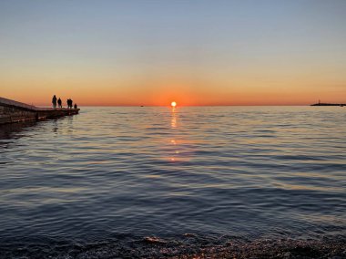 Aralık 'ta denizde gün batımı, berrak turuncu gökyüzü, mavi deniz, ufukta gemiler, dalgakırandaki insanlar gün batımını görüyorlar.