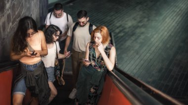 Bir grup arkadaş yürüyen merdivende dururken akıllı telefonlardan konuşur. Dışarıda cep telefonu olan modern insanlar. Mobil iletişim teknolojisi. Yüksek açı görünümü