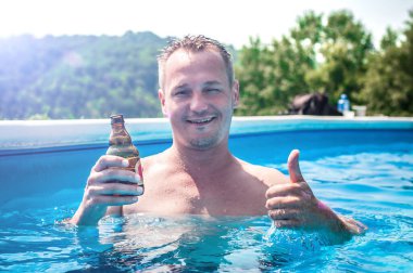 Şişme yüzüklü mutlu genç adam bira içer, dinlenir ve yazın havuzda eğlenir.