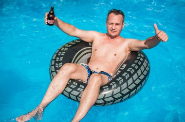 Şişme yüzüklü mutlu genç adam bira içer, dinlenir ve yazın havuzda eğlenir.