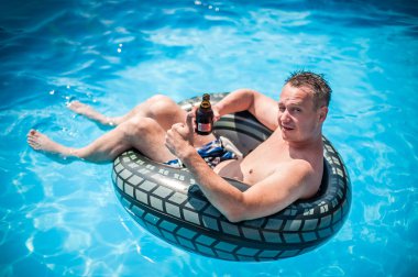 Şişme yüzüklü mutlu genç adam bira içer, dinlenir ve yazın havuzda eğlenir.