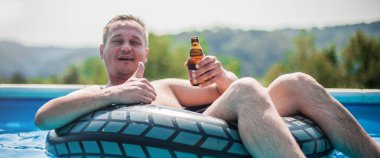 Şişme yüzüklü mutlu genç adam bira içer, dinlenir ve yazın havuzda eğlenir.