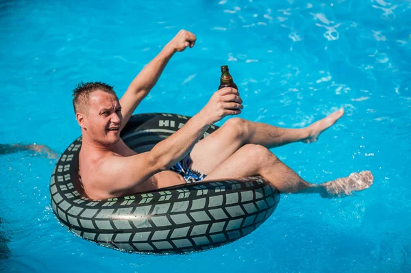 Şişme yüzüklü mutlu genç adam bira içer, dinlenir ve yazın havuzda eğlenir.
