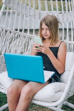 Dizüstü bilgisayar ve akıllı telefon kullanarak sallanan sandalyede oturan güzel odaklı kızın portresi. Açık havada boş zaman