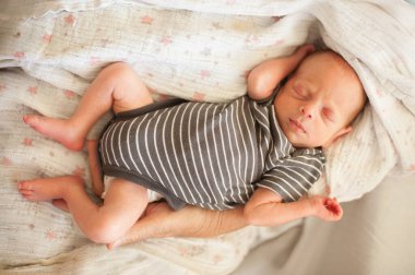 Küçük bir bebek, yumuşak bir battaniyeye sarılmış, mışıl mışıl uyuyor. Bebek, çizgili bir tulum giyiyor, huzurlu, sevgi dolu bir kucaklama içinde, iç açıcı bir sahne yaratıyor..