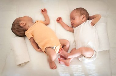 İki bebek rahat bir kreşte huzur içinde dinleniyor. Biri sarı tulum giyerken diğeri beyaz giyer. Yumuşak bir battaniyeye konforlu bir şekilde yerleştirilmişler, sakince yayılıyorlar..