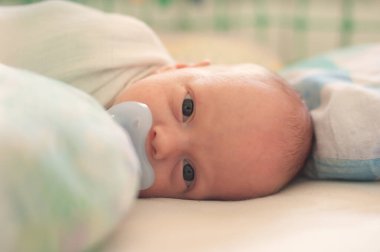 Küçük bir bebek, ağzında bir emzik ile bir beşikte sırt üstü yatıyor. Bebek uyanık ve doğrudan kameraya bakıyor. Huzurlu bir ifadeyle..