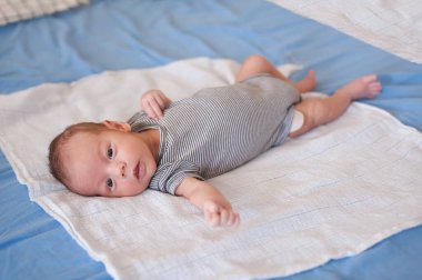 Çizgili tulumlu bir bebek yumuşak bir battaniyenin üzerinde merakla uzanır. Oda sakin ve parlak. Ufaklık için huzurlu bir ortam yaratıyor..