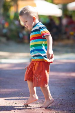 Genç bir çocuk, çizgili bir gömlek ve şort giyerek güneşin altında canlı bir oyun alanını keşfediyor. Sıcak zeminde çıplak ayakla mutlu bir şekilde yürüyor, maceralı gününün tadını çıkarıyor..