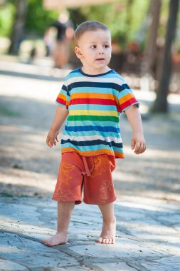 Genç bir çocuk parlak çizgili gömlek ve turuncu şort giyerek güneşli bir yolda çıplak ayakla yürüyor. Çevre ağaçlarla ve arka plandaki insanlarla dolu..