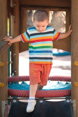 Genç bir çocuk bir oyun alanı yapısında dikkatlice geziniyor. Renkli çizgili bir gömlek ve turuncu bir şort giyiyor. Açık havada arkadaşlarıyla eğleniyor..