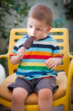 Küçük bir çocuk bahçedeki parlak sarı bir sandalyede oturur ve mutlu bir şekilde çikolatalı dondurma yer. Güneş parlıyor ve yeşil alan çevreyi sarıp neşeli bir atmosfer yaratıyor..