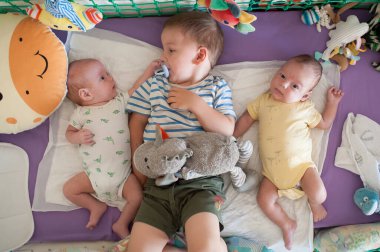 Üç küçük kardeş canlı bir kreşte oynuyorlar. En büyük kardeş oyuncak tutarken, küçük ikizler merakla izliyor. Yumuşak yatak ve neşeli oyuncaklar çevrelerini sarmış.