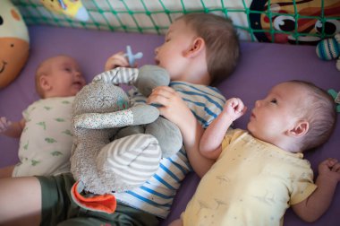 Üç kardeş oyun alanında birlikte yatıyorlar. Büyük kardeş bir oyuncak tutarken, ikizler onun yanında dinleniyor ve eğleniyorlar..