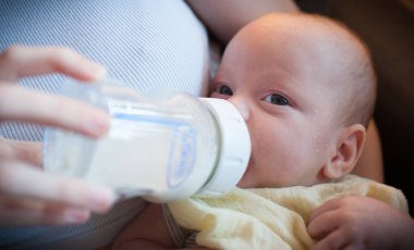 Bir bebek ebeveyninin kucağına rahatça oturur ve öğleden sonra bir şişeden su içer. Bebek beslenme saatine odaklanırken ebeveyn şişeyi tutar..