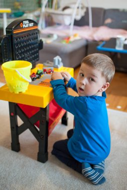 Küçük bir çocuk renkli bir alet takımıyla yerde oturuyor. Yakınlarda bir kova duruyor ve oda oyuncaklarla dolu. Güneş ışığı pencereden sahneye doğru geliyor..