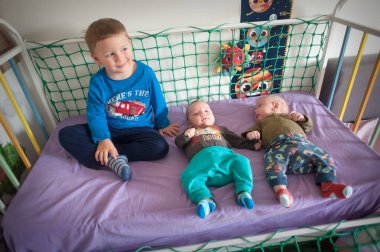 Ağabeyi beyaz bir beşikte lavanta renkli bir yatakta oturur ve küçük ikiz kardeşlerine bakar. Bebekler uzanmış, çorap ve pantolon giyiyorlar..