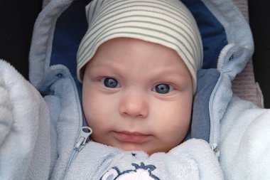 Mavi gözlü bir bebek bebek arabasında otururken doğrudan kameraya bakar. Bebek açık mavi bir elbise ve çizgili bir şapka giyiyor..