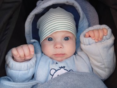 Mavi gözlü, açık mavi elbiseli ve çizgili şapkalı bir bebek bebek arabasında oturuyor. Bebek meraklı görünüyor ve gün boyunca ellerini içeride tutuyor..