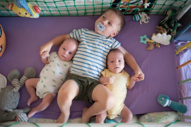 Üç küçük çocuk bir beşikte. Emziği olan bir ağabey, iki küçük ikiz kardeşiyle mor bir yatakta el ele tutuşur..