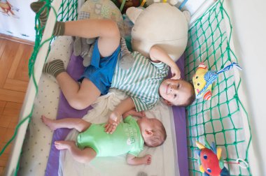 Şort ve çizgili gömlek giyen genç bir çocuk oyuncak ve peluş hayvanlarla dolu bir oyun parkında küçük kardeşinin yanında yatıyor. Bebek yeşil bir tulum giyiyor..