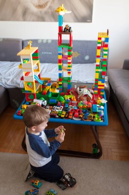 Genç çocuk yerde diz çöküyor ve küçük bir oyuncak arabayla oynuyor. Arkasında daha çok oyuncak araba ve yüksek bina blokları olan bir masa var..