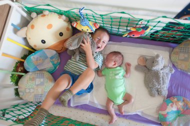 Bir ağabey, beşiğinde kardeşinin yanında gülerken oyuncak bir hayvana sarılır. Etraflarını saran renkli oyuncaklar ve pencereden içeri güneş ışığı giriyor..