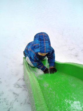 Bir çocuk oyun parkında karla kaplı yeşil bir slayta tırmanıyor. Çocuk mavi bir ceket giyiyor ve karın içine doğru kaymaya odaklanıyor..