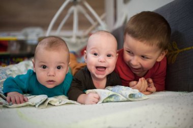 Üç erkek kardeş birlikte zaman geçiriyorlar. Bir ağabey, iki küçük ikiz kardeşine karnının üzerinde battaniyeye uzanmış şekilde gülümsüyor..