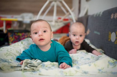 İki küçük ikiz kardeş, biri mavi gömlekli, diğeri koyu renk gömlekli, oyun odasında çiçeklerle süslü bir battaniyenin üzerinde yatıyorlar..
