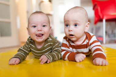 İki sevimli bebek ikiz kardeş sarı bir paspasın üzerinde yan yana yatıyorlar. Bir kardeş dili açık gülümserken, diğeri yüzünde şaşkın bir ifadeyle yukarı bakıyor..