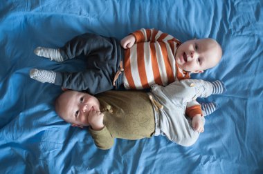 İki sevimli ikiz kardeş mavi bir çarşafın üzerinde yatıyor. Bir kardeş parmaklarını emerken diğer kardeş yukarı bakıyor ve gülümsüyor. Birlikte vakit geçiriyorlar..