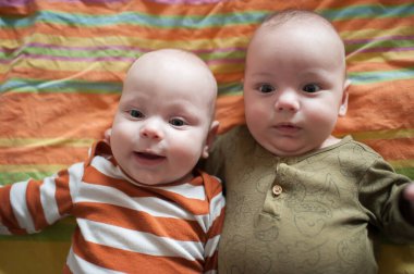 İki mutlu ikiz kardeş renkli bir battaniyenin üzerinde yan yana uzanıyorlar. Bir bebek gülümserken diğeri meraklı görünüyor. Birbirlerine çok yakınlar ve kucaklaşıyorlar..