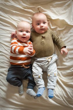 İki bebek yan yana bir yatakta yatıyor. Gülümsüyorlar ve rahat kıyafetler giyiyorlar. Yumuşak yatak, oynamak için sıcak bir ortam yaratır..