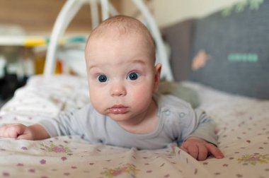 Bir bebek yatağın üzerinde karnının üstünde dinleniyor. Bebeğin büyük gözleri var ve merakla kameraya bakıyor. Ayar yumuşak renklerle aydınlık..