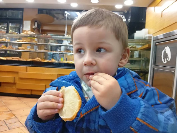 Küçük bir çocuk pastanede elinde pastayla oturuyor. O, aperatifini tadarken meraklı görünüyor. Dükkanın arka planında çeşitli fırınlanmış ürünler sergileniyor..