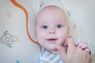 Parlak gözlü bir bebek, yumuşak bir yüzeyde yatarken bakıcıya gülümser. Sabahın ilk ışıklarındaki ortam sıcak bir atmosfer yaratıyor..