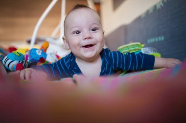 Sabah ışığında renkli oyuncaklarla çevrili yerde oynarken neşeli bebek gülümsüyor. Çizgili gömlek giyen bir bebek, karnının üstüne yatar, kollarıyla desteklenir..