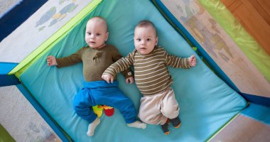 İkiz erkek kardeşler, parlak renkler ve oyuncaklarla çevrili yumuşak bir oyun hasırının üzerinde yatıyorlar. Onların masum gülümsemeleri ve tuhaf ifadeleri bu rahat yerde çocukluk sevincini yakalar..